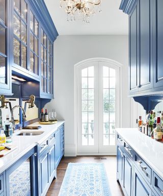 A blue galley kitchen with painted cabinets, white walls and worktops, chandelier ceiling light and runner rug