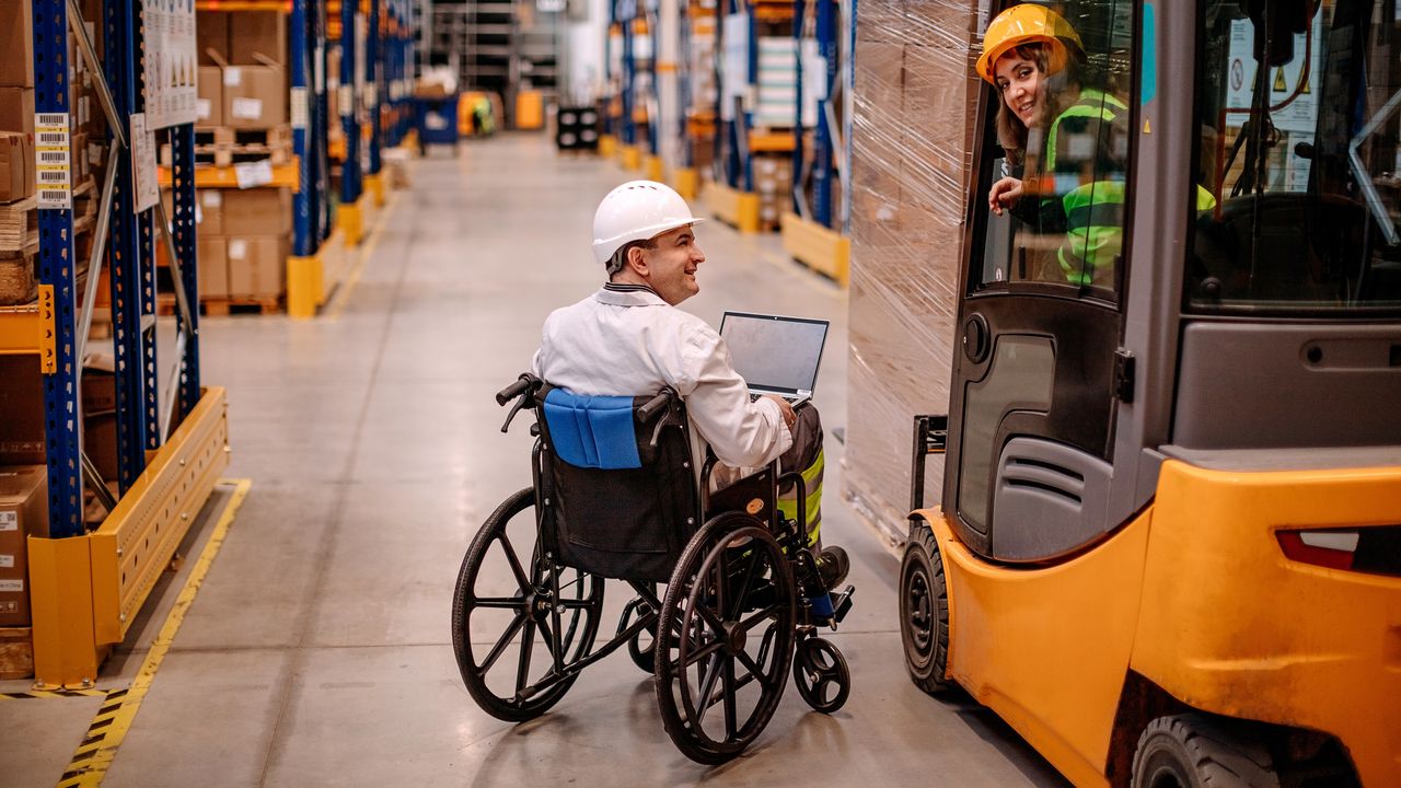 A male worker wearing a hard hat and in a wheelchair speaks with a female worker on a forklift in a warehouse.