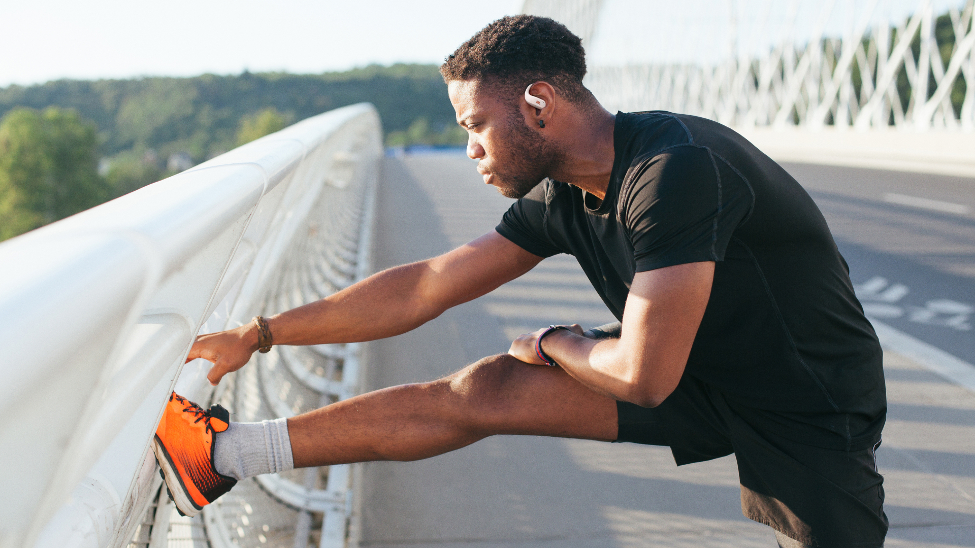 JBL Endurance Zone worn by a male runner, stretching his hamstrings on a suspension bridge, on a sunny morning