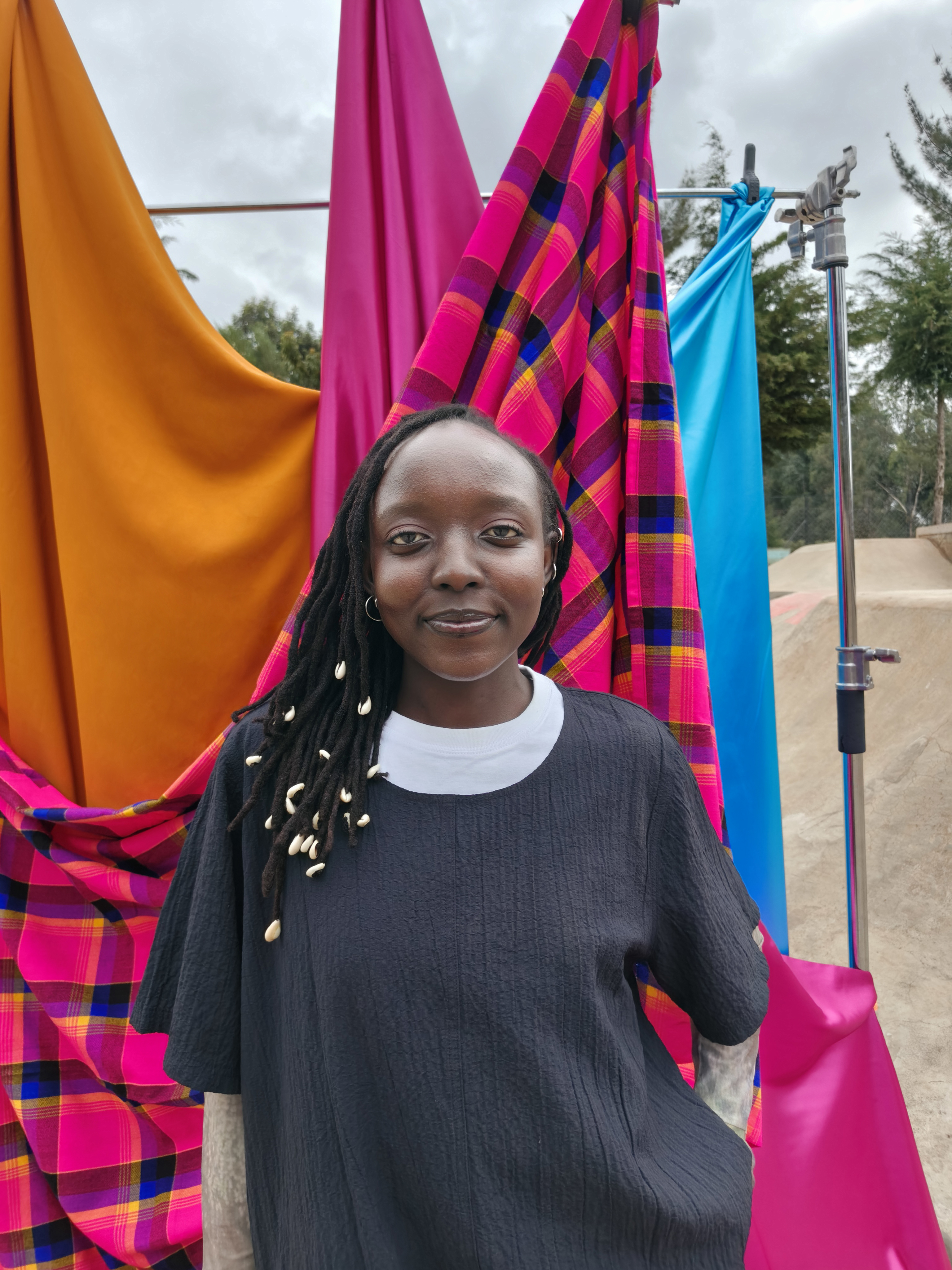 A woman with locks threaded with cowrie shells, wearing a black textured top over a white T-shirt, stands in front of orange, pink, checked pink and turquoise fabrics hung on a stand at a skatepark.