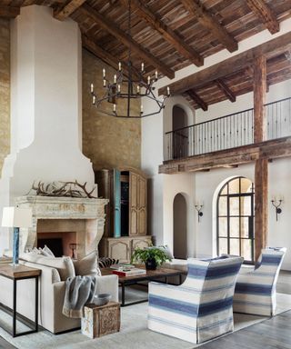 Living room with grand double height ceiling, dark wooden ceiling, large stone chimney and fireplace, wooden balcony, seating area around fireplace, two striped armchairs, cream sofa, dark wood and metal coffee table, black metal chandelier