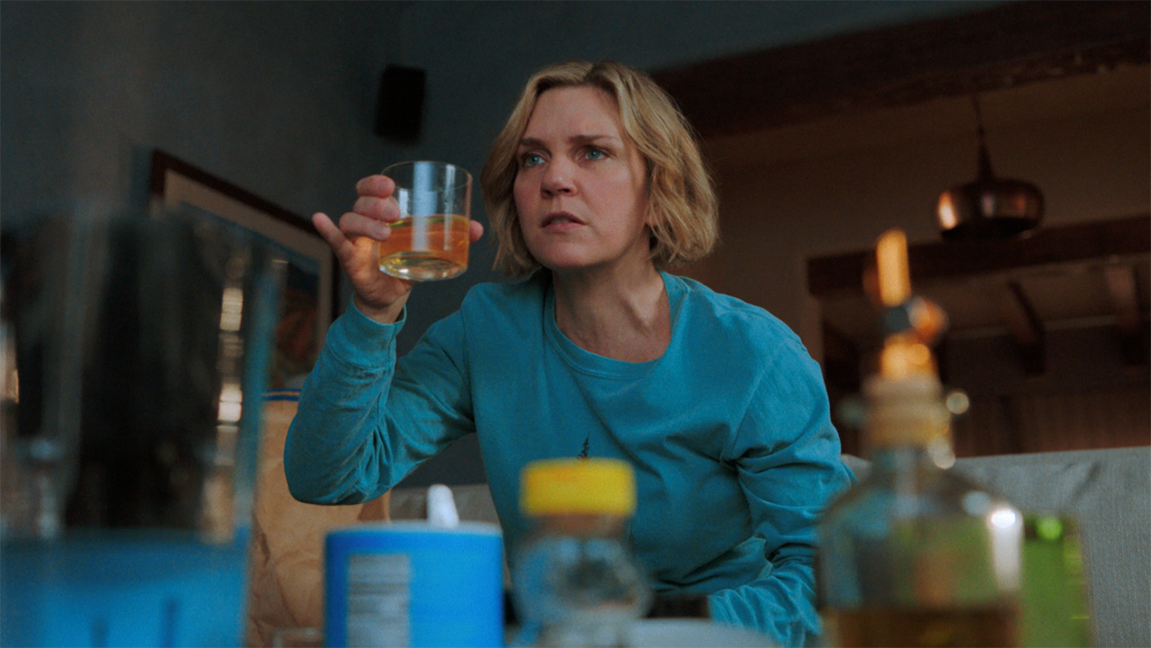 Carol sitting down and holding up a glass of orange liquid in Pluribus episode 5
