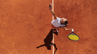 Female tennis player is shown serving a tennis ball on an orange court. She is stood in the center of the image by a white line on the court.