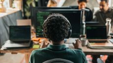 AI coding concept image showing female software developer wearing headphones while working on a desktop computer in an open plan office space. 