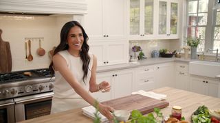 Meghan Markle cooking in a white kitchen and smiling wearing an ivory sleeveless sweater and white skirt