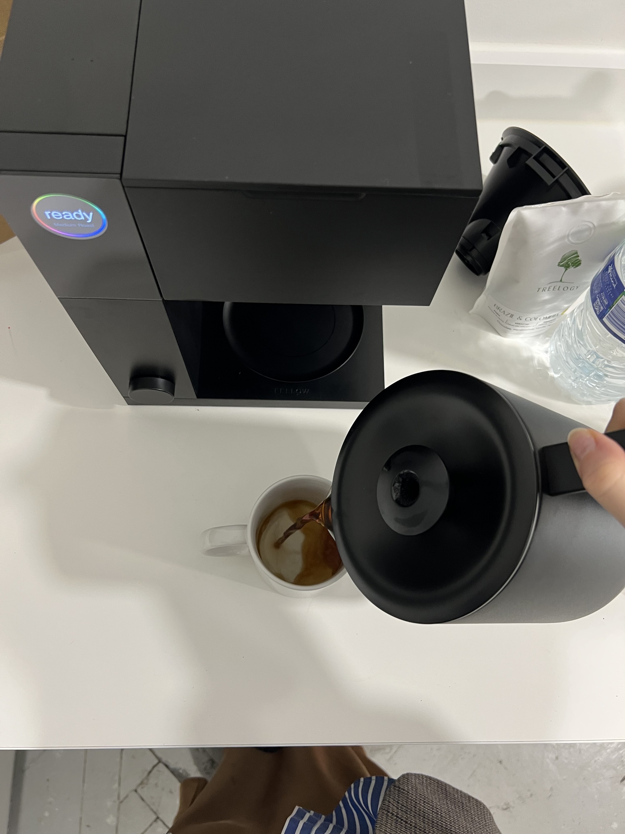 Image of a hand pouring coffee from a black coffee pot into a white mug on a white kitchen countertop with the black Fellow Aiden Precision coffee maker in view.