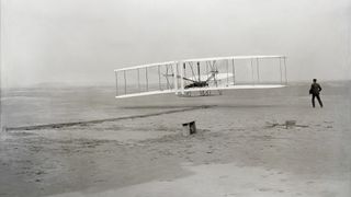 The first powered, controlled, sustained airplane flight in history. Orville Wright, age 32, is at the controls of the machine, lying prone on the lower wing with hips in the cradle which operated the wing-warping mechanism. His brother, Wilbur Wright, age 36, ran alongside to help balance the machine, having just released his hold on the forward upright of the right wing. The starting rail, the wing-rest, a coil box, and other items needed for flight preparation are visible behind the machine.