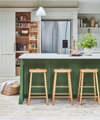 A kitchen with a moss green kitchen island with a white countertop and three light wooden stools, and light gray cabinets and a silver fridge in front of it