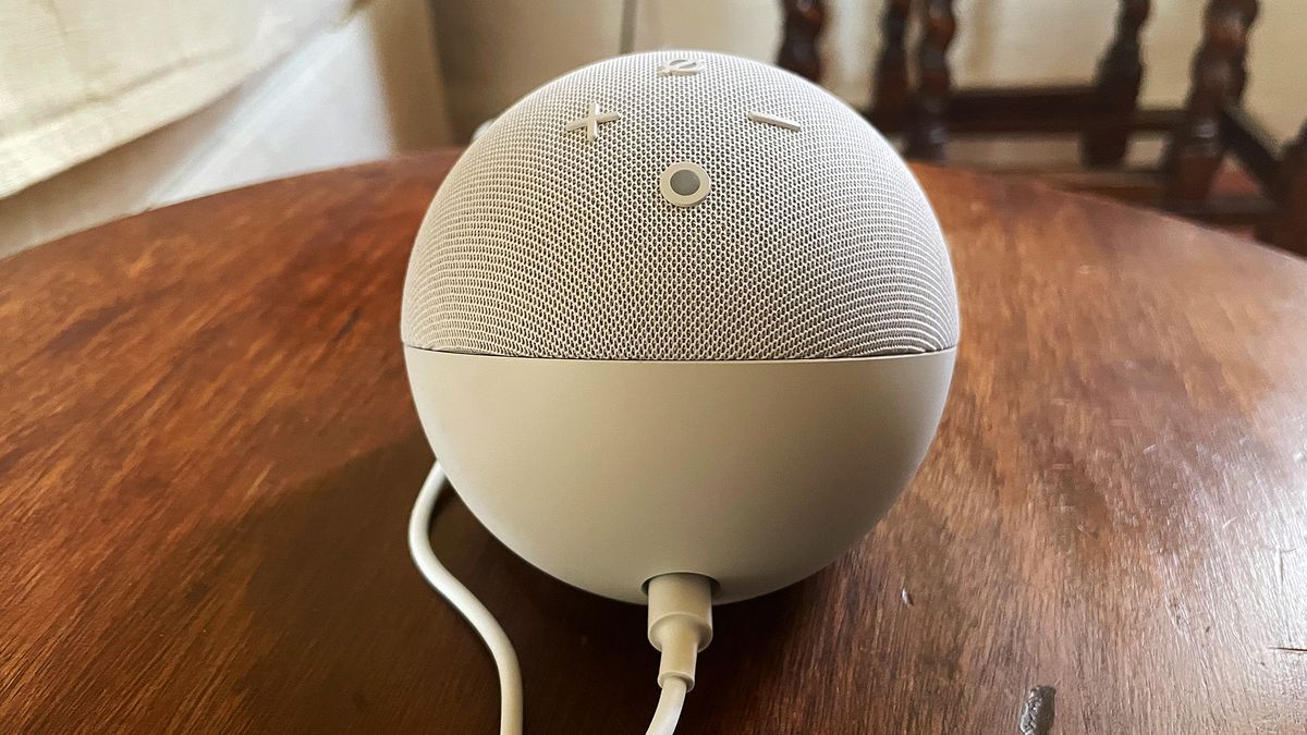 The image shows the rear of a white Amazon Echo Dot (5th Generation) smart speaker. It is round and there are four buttons visible on top. The power cable protrudes from underneath.
