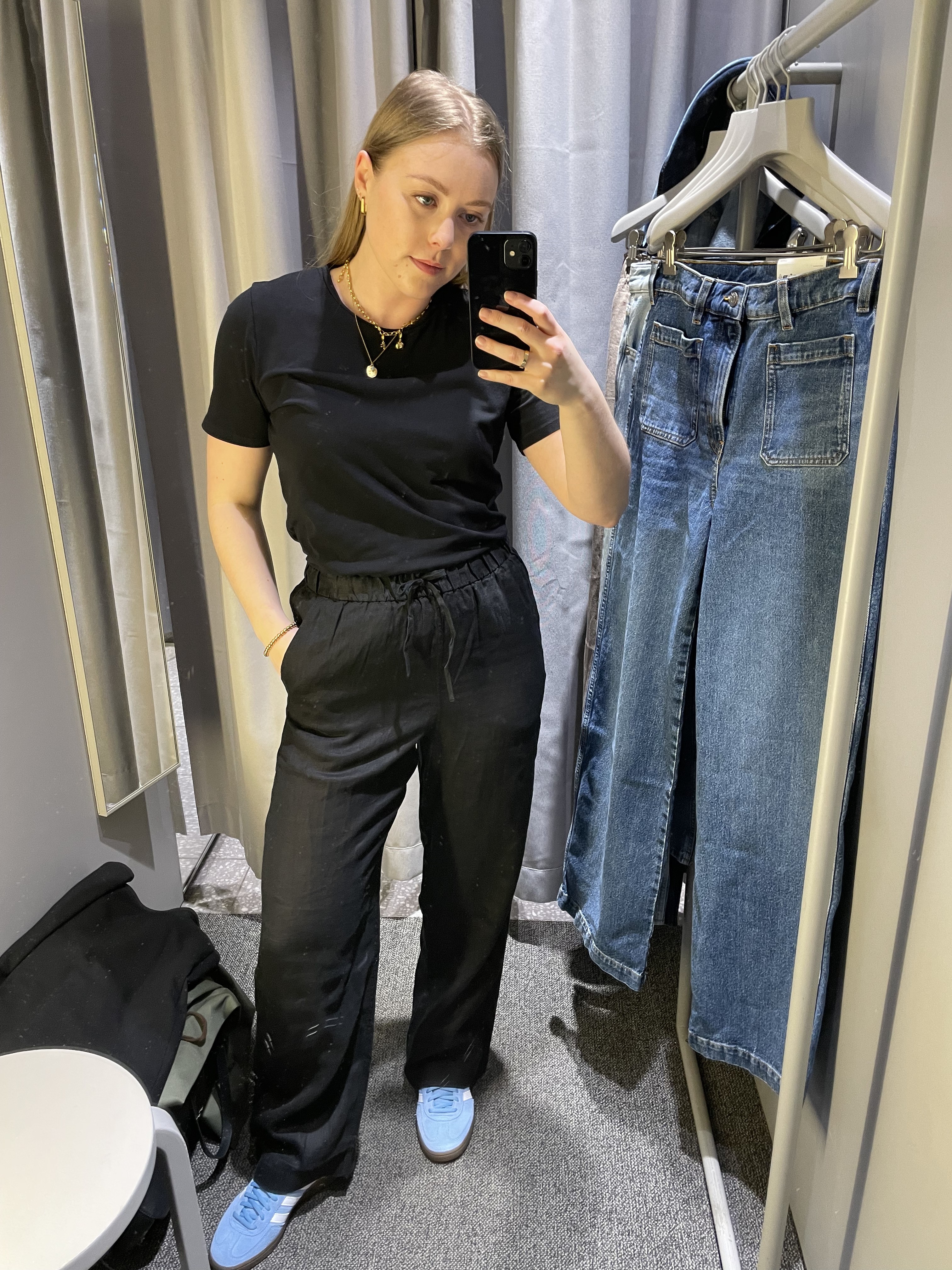Woman in dressing room wears black t-shirt, linen trousers, blue trainers