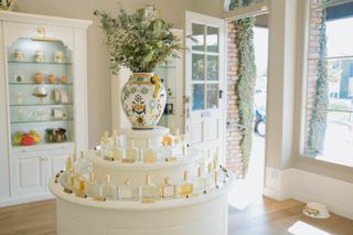 A perfume shop features traditional interior decor in the tones of white, cream, light blue, and brown. Decorations include a wide, floral design vase in yellow, white, and blue, and numerous perfume bottles sitting on a central pillar as well as on a shelving unit at the bottom of the room.