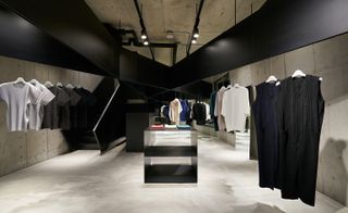 A concrete space with black wide beams across the roof from which the sportswear-inspired clothes are neatly hung and a glass counter in the middle of the room with a staircase going up one side of the room.