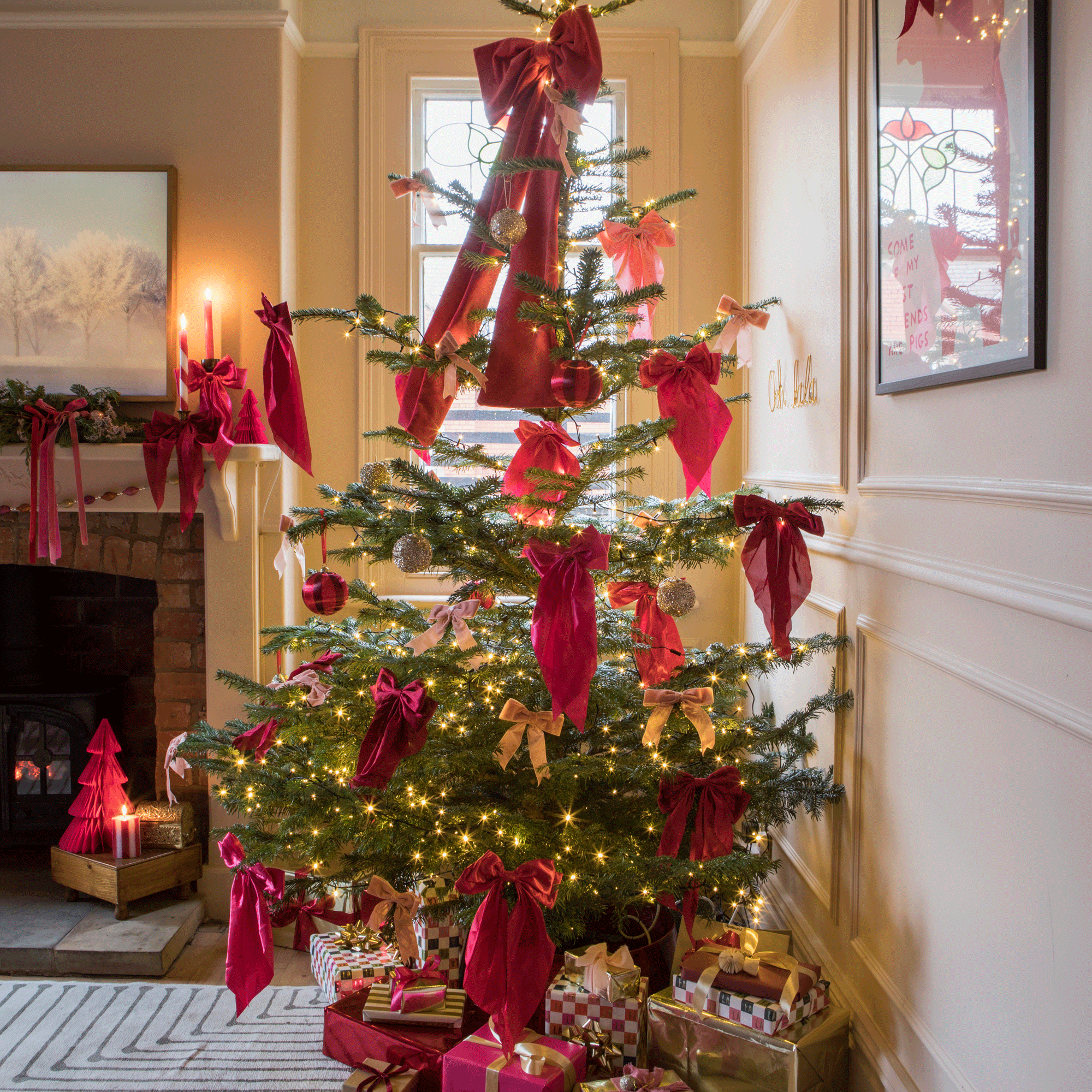a living room with a Christmas tree covered in large pink/red bows, a mantelpiece with festive greenery, bold typography artwork