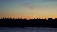 A crescent moon shines in the evening sky above a forest as two planets glow overhead in the peach colored sky.