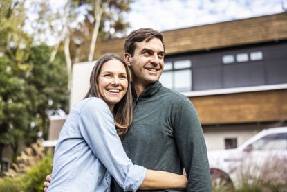 Portrait of married couple in front of modern home. They are in their late-30s or early-40s.