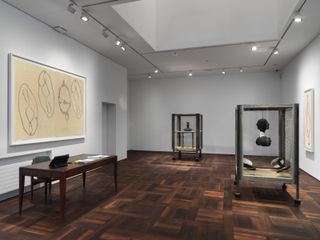 An art gallery featuring white walls and ceilings , white ceiling lights and dark wooden floors. Sculptures in glass museum display case. Wall art on display. Dark brown table with chair in the left of the room