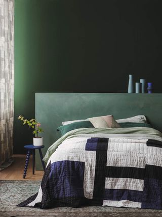 Image of a bed with green, blue, and white bedding. There is a sage green, extended headboard behind it and the walls are a darker shade of green.