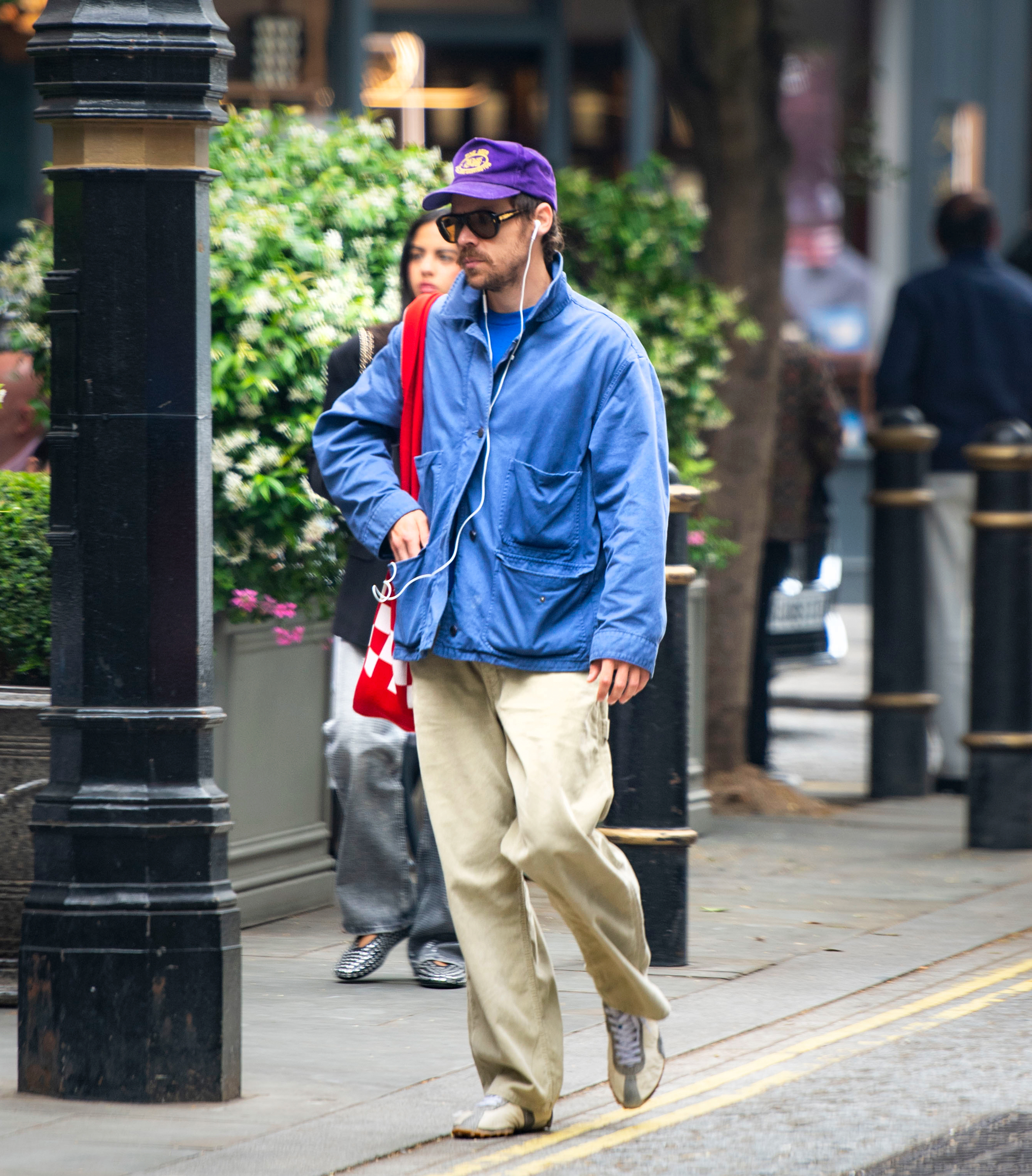 Harry Styled walks down the streets wearing a cobalt blue chore jacket, beige trousers, a purple baseball cap and Maison Margiela Sprinter trainers.