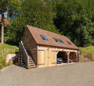 An oak frame triple bay garage with clay roof tiles and rooflights in the roof serving a room above. The garage is set on a rural drive with trees in the background.