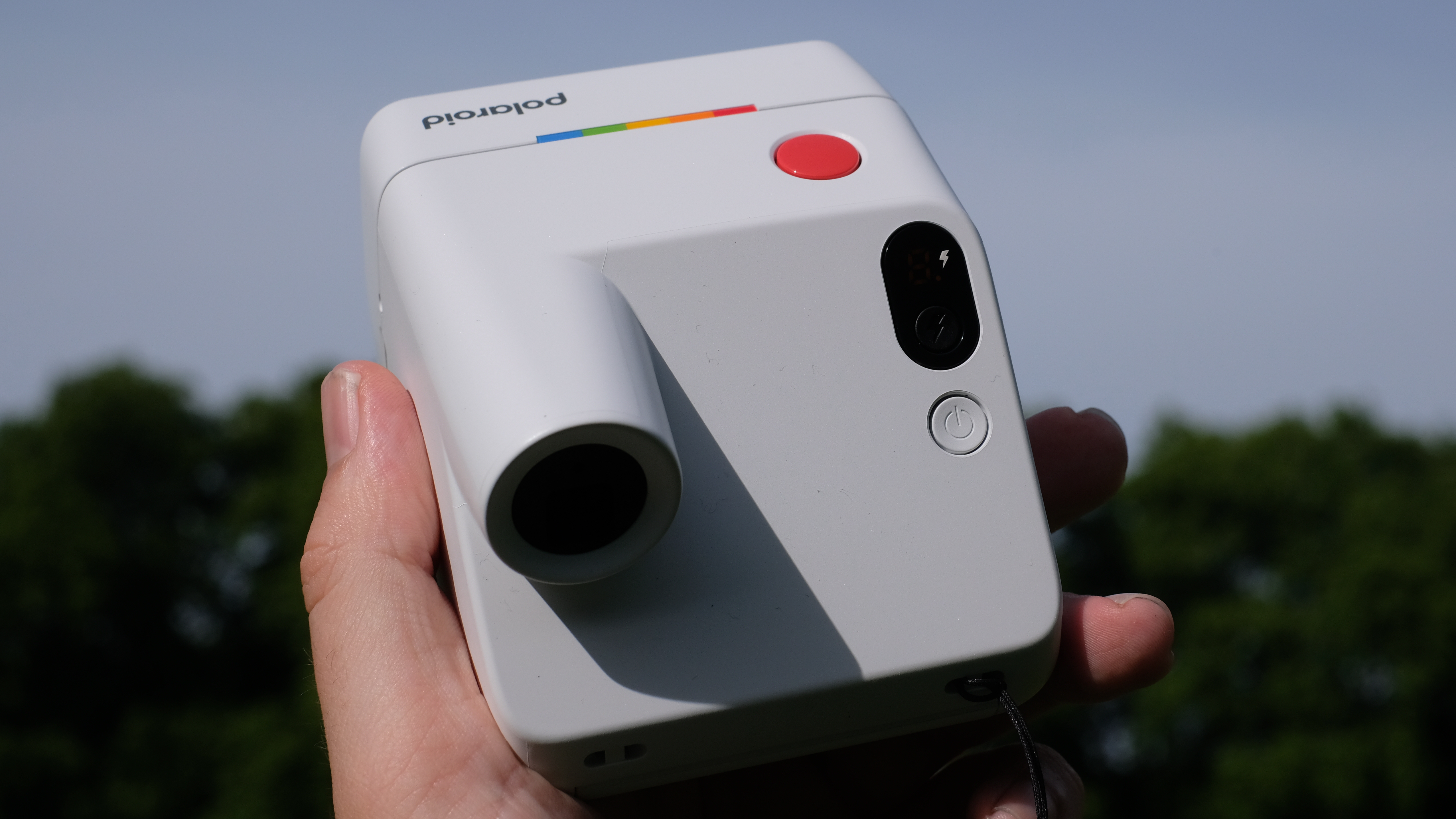 Photograph of man's hand holding Polaroid Go Gen 3 camera in white, in dramatic sunshine with significant shadows, pointing upwards and showing raised viewfinder and rear power button