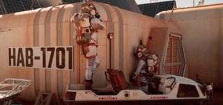 two people in spacesuits work outside a simulated mars habitat
