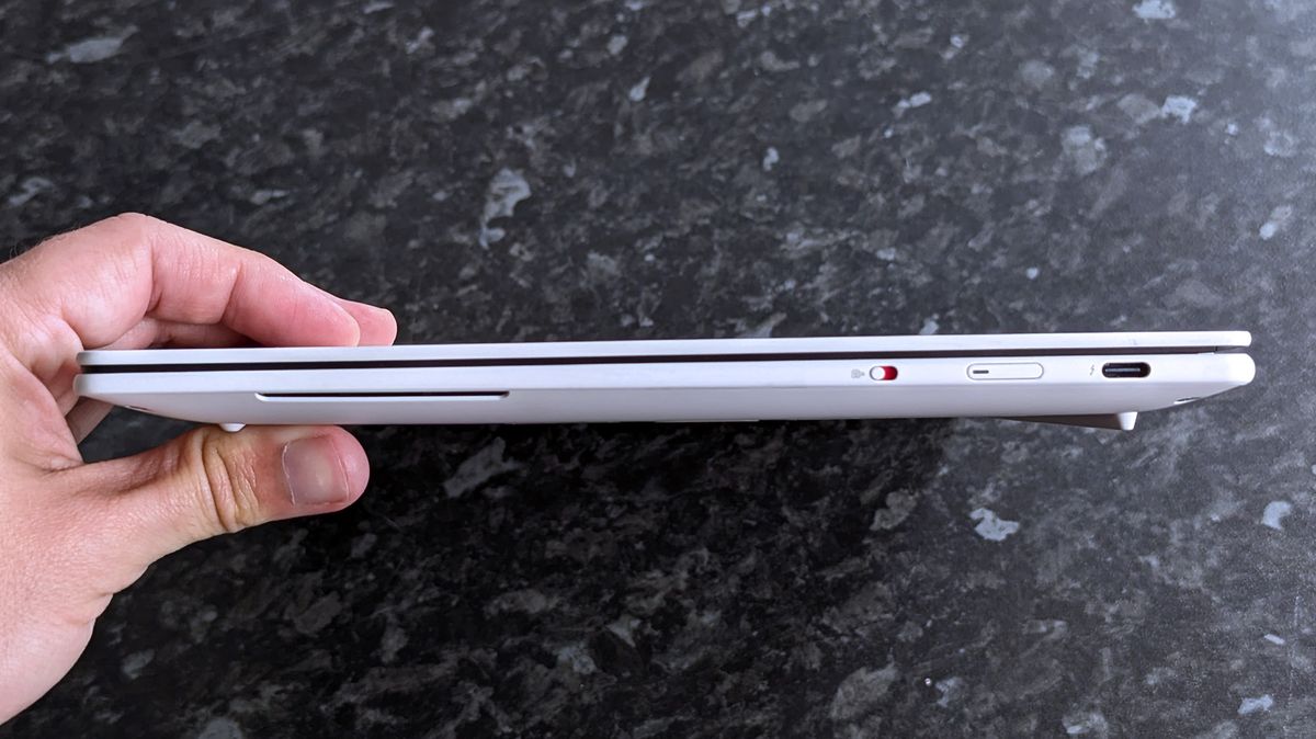 A hand holds a slim Lenovo Yoga Slim 7i Ultra (Gen 11) Aura Edition laptop horizontally over a dark, textured surface. The device features a slot, a red switch, and a USB-C port.
