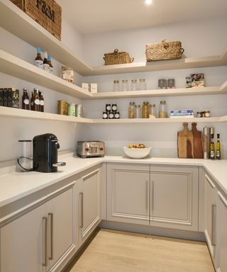 pantry room with light wooden floor, pale taupe coloured base units, white worktop and three sets of shelving running around top section of walls