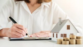 Woman signing document next to a model of a house and stacked coins.