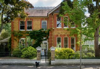 exterior of a large London Victorian home with original conservatory, with a black gate surrounding, trees, and a dog at the front