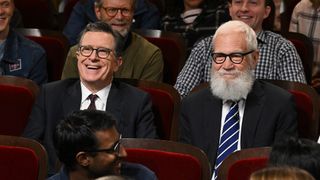 A late night still of Stephen Colbert -- both in suits, ties, and glasses -- sitting in the audience at The Late Show.