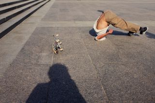 Daytime image of male skateboarder in a falling position on a concrete floor, skateboard and stone steps to the left , shadow of photographer front shot