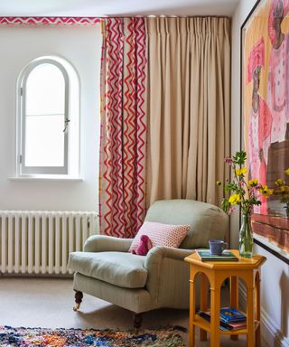 Girl's bedroom with pink patterned and cream curtains, small arched window, beige cozy armchair, cream carpet, colorful wall art, bright yellow side table