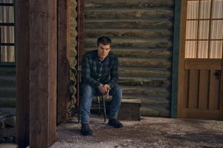 Michael C. Hall in a plaid shirt and jeans sitting outside a cabin