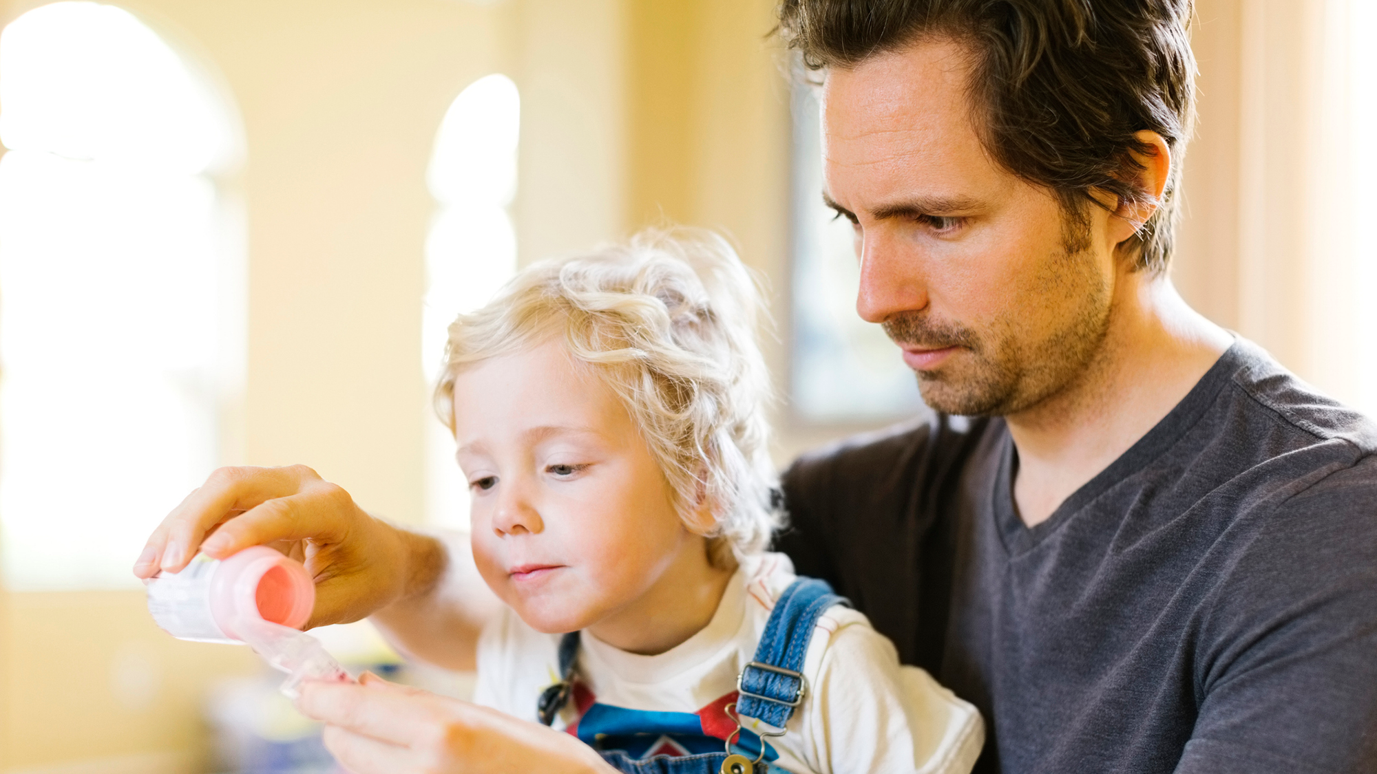 A blond child wearing overalls sits on the lap of a dark haired man wearing a gray shirt pouring something for the child.