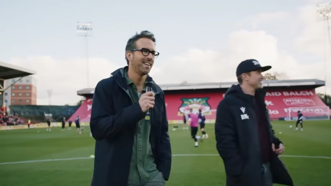 Ryan Reynolds talking into a microphone on a football pitch with Rob Mc Elhenney standing behind him in Welcome to Wrexham.