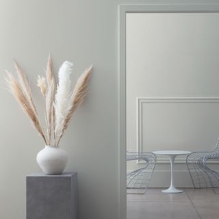 light gray wall with plinth with pampas leaves, doorway leading into gray room with metal chairs and round white table