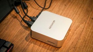 A compact Geekom mini PC sits on a wooden surface connected to several cables. It has a sleek, modern design with a metallic finish, conveying a tech-savvy vibe.
