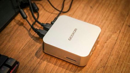 A compact Geekom mini PC sits on a wooden surface connected to several cables. It has a sleek, modern design with a metallic finish, conveying a tech-savvy vibe.
