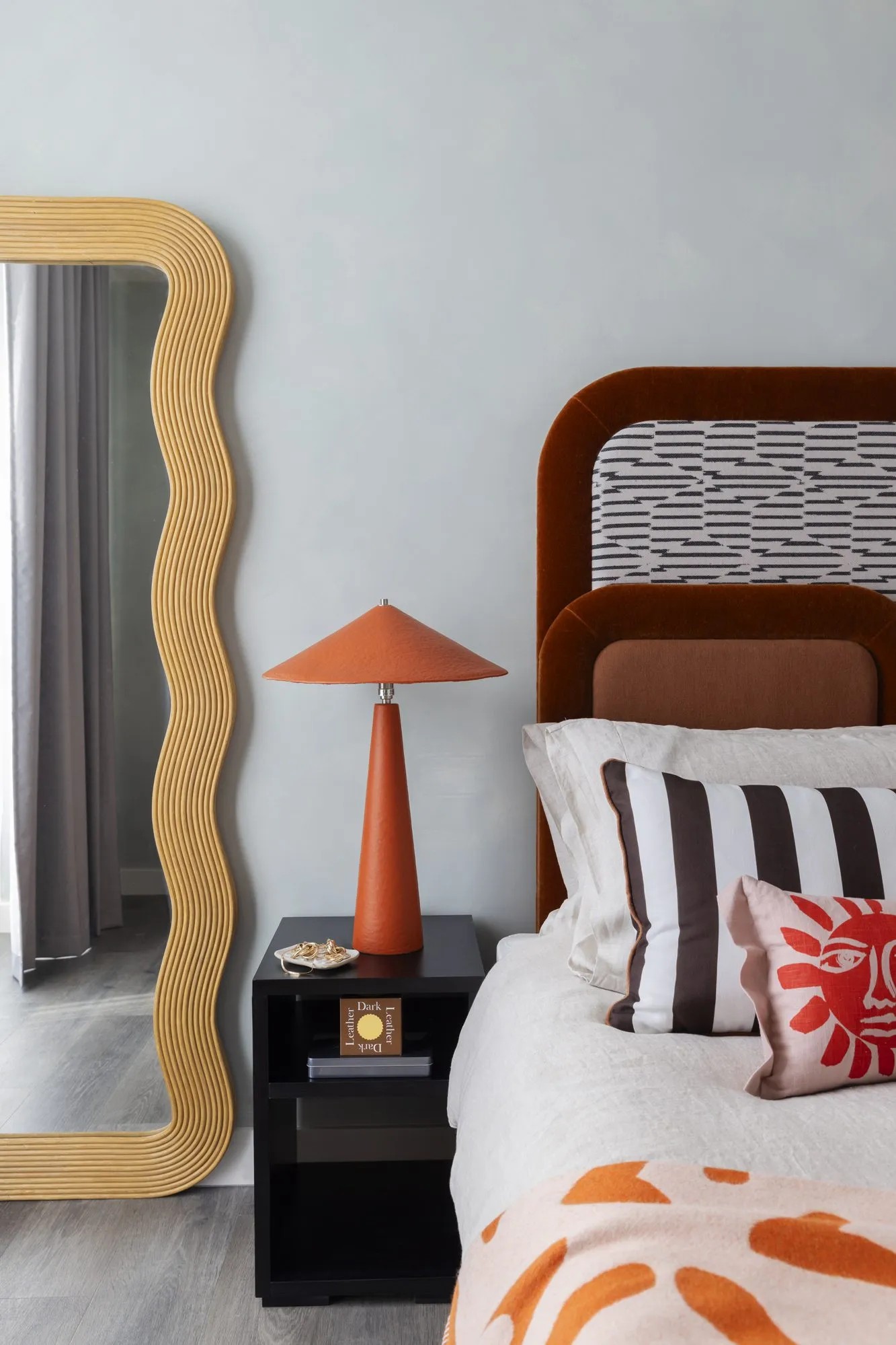 A bedroom with a wavy wood mirror, a terracotta tinted bedside table lamp on a black bedside table, stripy cushions and patterned ones on a bed with a brown heaboard in velvet.