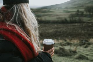 A girl looking out at a country side view while wearing a black hat and a red scarf, and holding a takeaway coffee cup.
