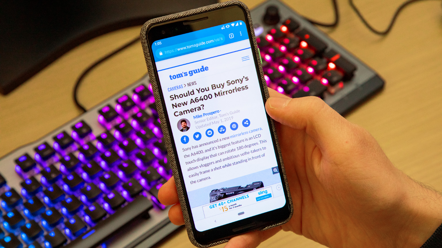  A hand holding a black Android smartphone with the Tom's Guide website open on the screen and a backlit mechanical keyboard in the background.