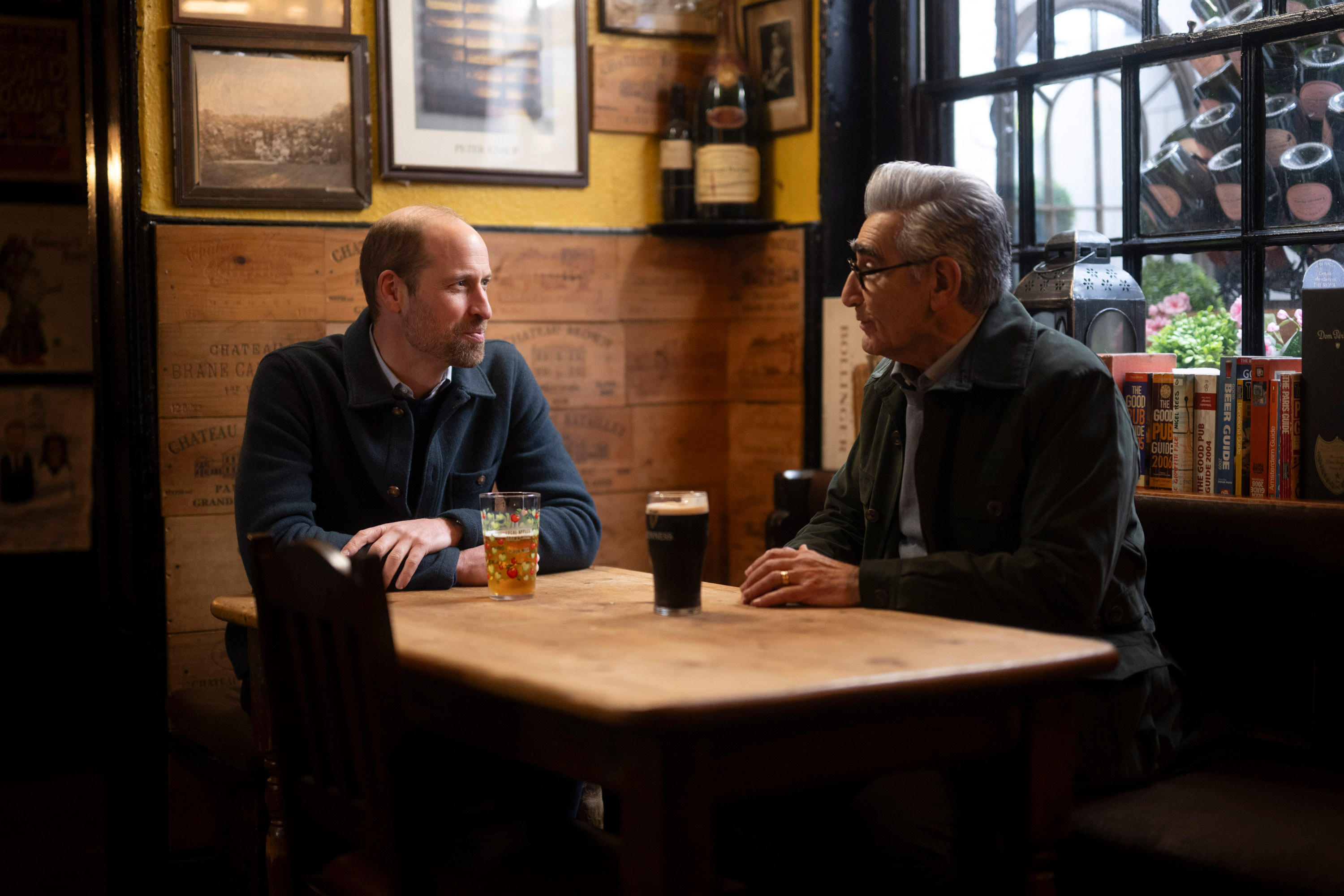 Prince William and Eugene Levy drinking beers at at a table in a pub