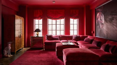 Image of a red color-drenched lounge room with three windows that have red curtains, a burl cabinet, and a giant red sectional sofa.