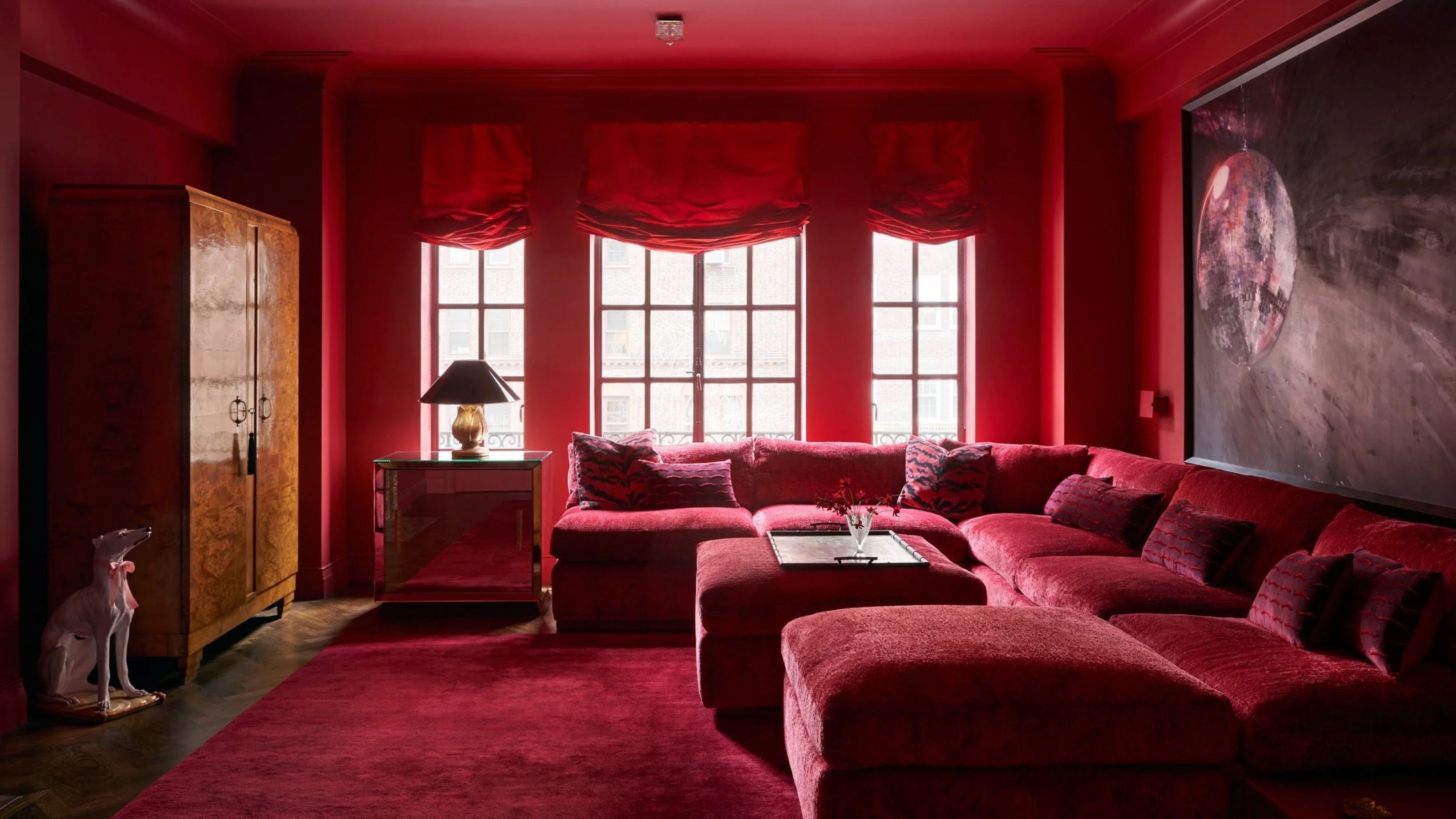 Image of a red color-drenched lounge room with three windows that have red curtains, a burl cabinet, and a giant red sectional sofa.