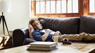 A man in a blue shirt takes a nap after reading on his brown leather couch