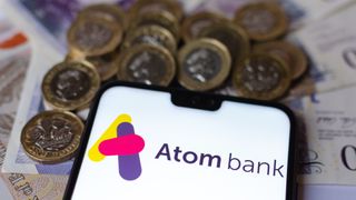 Atom bank app logo appearing on a smartphone which is sat on top of UK bank notes, surrounded by one-pound coins