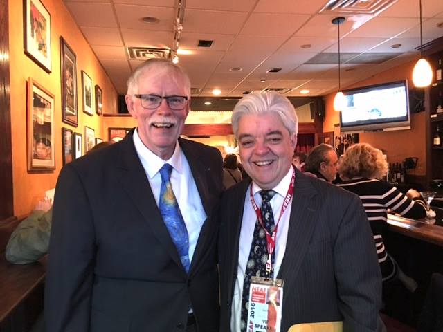 two men in suits smile for a photo in a cafe