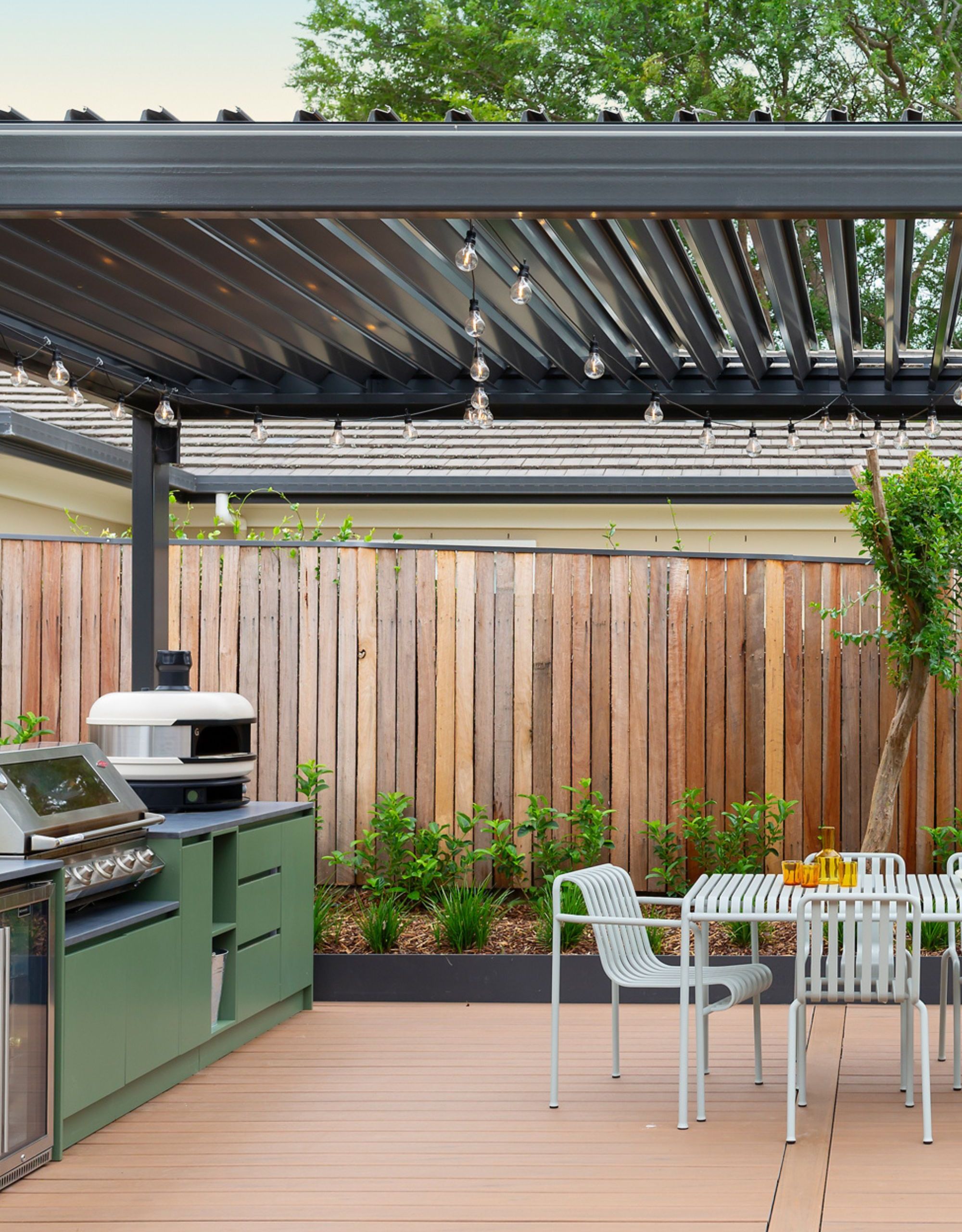 A large pergola above an outdoor kitchen with green cabinetry and a pizza oven and a dining area with HAY-like metal table and chairs