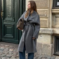 Influencer @claradyrhauge wears a grey wrap coat with blue jeans and black kitten heel boots.