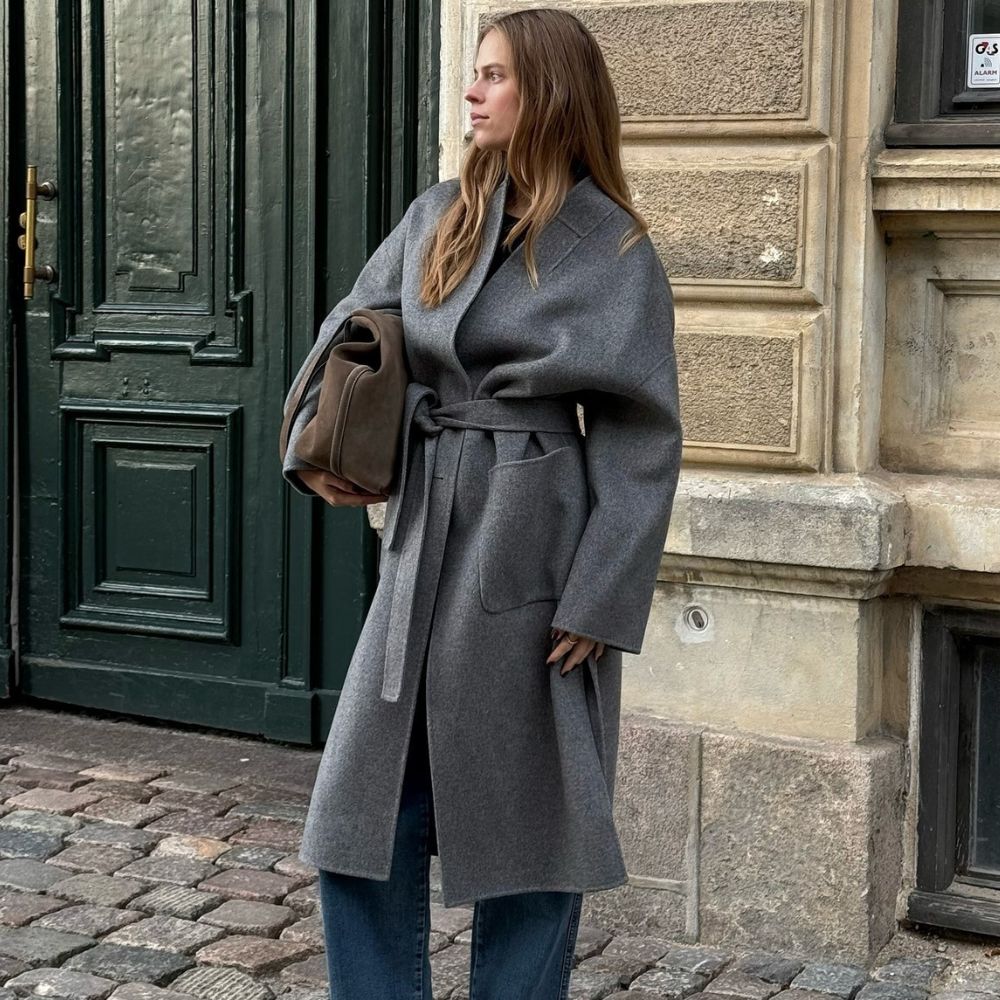 Influencer @claradyrhauge wears a grey wrap coat with blue jeans and black kitten heel boots.